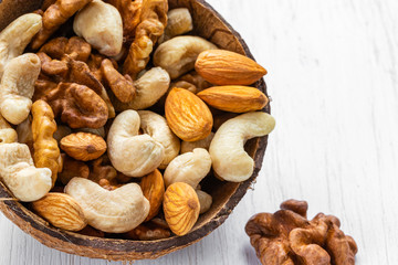 A mixture of nuts lies in a half of a coconut shell on a white wooden surface, top view