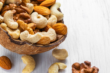A mixture of nuts lies in half a coconut shell and on a white wooden surface, side view from above
