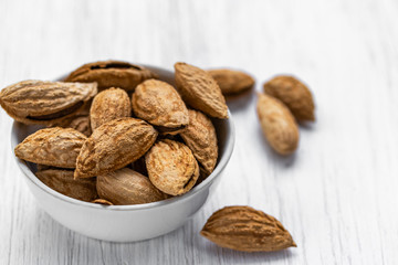 Inshell almonds lie in a white glass cup on a white wooden surface, side view from above
