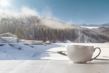 Single tea or coffee cup and landscape of mountains on background. Cup of hot drink with snowly look and cloudly sky in front of it. Warm in winter day, holidays, travel, New Year and Christmas time.