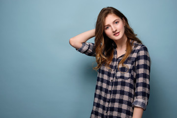 Obraz premium A young girl in a plaid shirt stands on a blue background and looks at the camera with her head bowed, her hand behind her head.