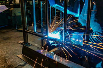 A welder at work in a workshop produces metal structures. Sparks from welding fly around.