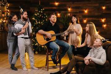 Handsome young man plays guitar and sings in front of friends at a Christmas party in a cozy house....