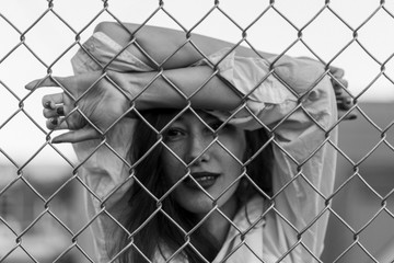 Pretty woman stands against wired fence. Model in white shirt. Purple lips. Pretty make up. Posing for magazine. Street fashion. Urban lifestyle. lady with long hair, beautiful eyes, behind fence.
