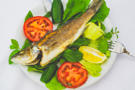 grilled trout and spinach, mint, greens, lemon, tomato and cucumber slices on the white plate with knife and fork on white background. Part of low-calorie diet