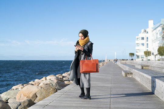 Woman wearing headscarf using cell phone by ocean with briefcase