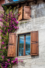 Fototapeta na wymiar Pink Bougainvillea Flowers on the Wall of Italian Stone House.Beautiful Facade of a House in Italy with Wooden Shutters on the Windows 