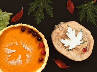 Homemade traditional american pumpkin pie on a black background. Decorated with powdered sugar in the form of a leaf and cranberries. Against the background of colorful autumn leaves. Top view.
