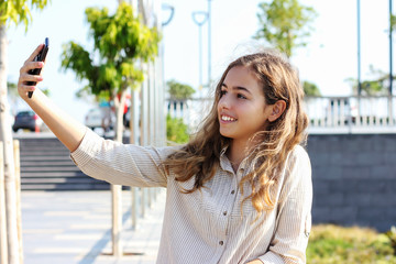 Beautiful teenager girl taking a selfie on the promenade on a sunny day. Happy young girl smiling with a phone in her hands