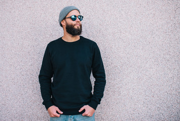 City portrait of handsome hipster man with beard wearing black blank hoody or sweatshirt and hat with space for your logo or design. Mockup for print