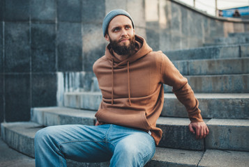 Obraz premium City portrait of handsome hipster guy with beard wearing brown blank hoodie or hoody and hat with space for your logo or design. Mockup for print