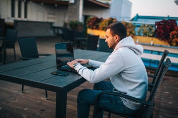 Young freelancer in gray hoodie uses laptop sits in cafe and reads news, checks e-mail. E-news concept.