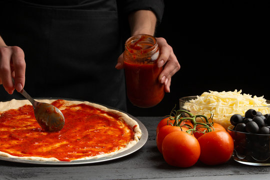 Chef smears Italian pizza with sauce, on black background, close-up. With ingredients mazzarella cheese, olives and tomato