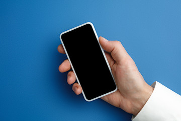 Male hand holding smartphone with empty screen on blue background for text or design. Blank gadget templates for contact or use in business. Finance, office, purchases. Mock up. Copyspace.