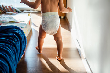 Baby in diapers learning to walk in her bedroom barefoot. - obrazy, fototapety, plakaty