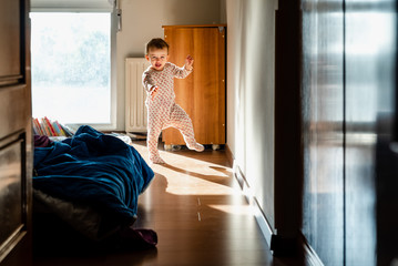 Newly awake baby in pajamas takes her first steps in the bedroom.