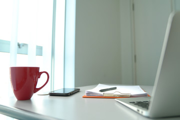 Office table next to windows. Computer, cell phone, coffee cup and focus on clipboard.