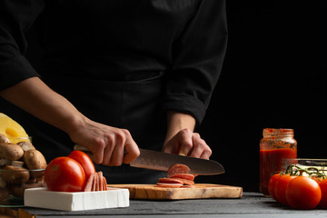 Chef prepares pizza, slicing fresh salami, on the background with ingredients. Recipe book, menu, home cooking.