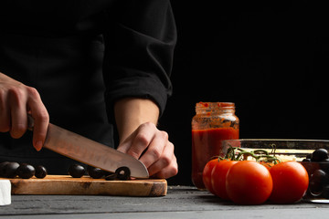 Chef prepares pizza, slices olives, on the background with ingredients. Recipe book, menu, home cooking, pizzeria