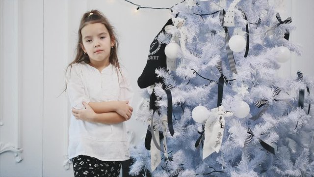 Bored little girl standing near fir-tree, frowning, looking very unhappy, because she doesn't like holidays, gifts, celebration and christmas.