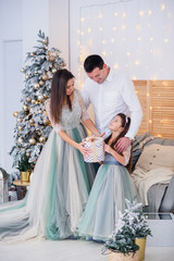 Happy girl unpacks holidays gift box with her parents while standing near Christmas tree. Happy lovely family.