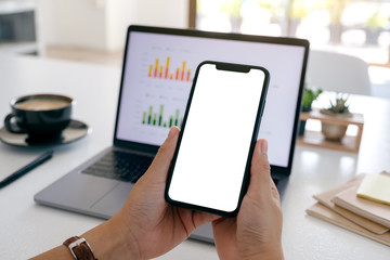 Mockup image of hands holding a black mobile phone with blank screen while using laptop computer in office
