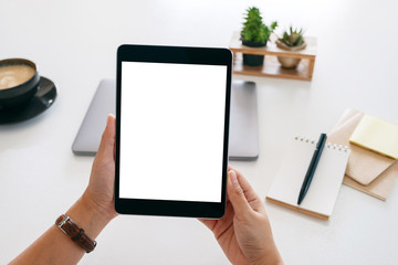 Mockup image of hands holding a tablet with blank white screen and laptop computer on wooden table...