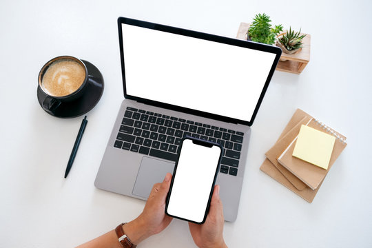 Top view mockup image of hands holding a blank white screen mobile phone and laptop computer on the table in office
