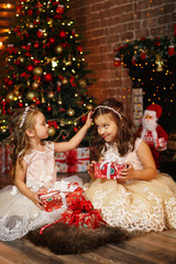 Little charming sisters in luxurious elegant dresses exchange gifts on Christmas Eve. Room in...