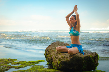 Fototapeta na wymiar Young woman sitting on the rock, meditating, practicing yoga and pranayama at the beach, Bali. Hands raising in namaste mudra