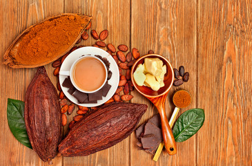 Cup of cacao drink with cocoa pods, cacao powder, cocoa butter, chocolate, sugar, cacao beans and natural green leaves on wooden background.