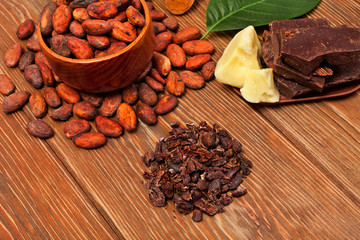 Cocoa beans in a spoon with green leaves, chopped chocolate, cocoa butter and heap of cocoa nibs on a wooden background.