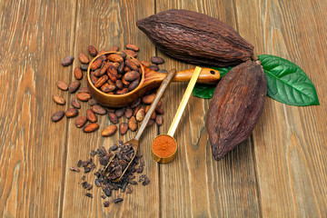 Cocoa beans in spoon, cocoa pods with green leaves, cacao powder in metal spoon and cocoa nibs in scoop on wooden background.