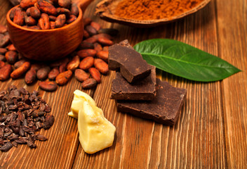 Cacao beans in wooden spoon with green leaves, chopped chocolate bar, cocoa butter, cacao powder...