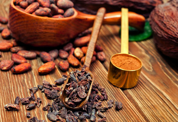 Cocoa beans in spoon, cocoa pods with green leaves, cacao powder in metal spoon and cocoa nibs in scoop on wooden background.