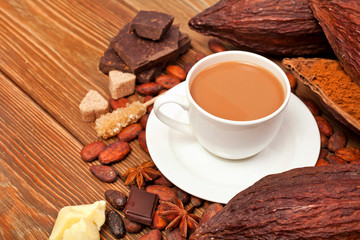 Cup of cacao drink with cocoa pods, cacao powder, cocoa butter, chocolate, sugar, cacao beans and star anise on wooden background.
