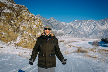 Fototapeta na wymiar Young cheerful guy enjoying Christmas vacation in mountains. Attractive man laughs and gives thumbs up against snow-capped mountains and blue sky. New year's journey, active winter tourism concept.