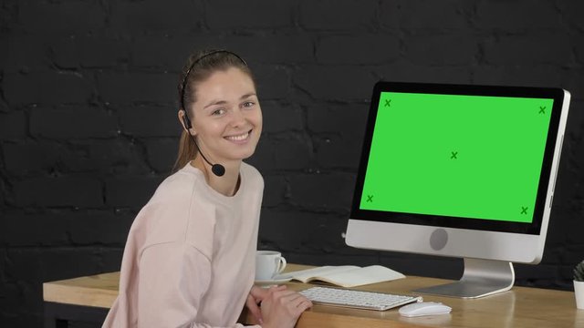 Smiling Woman Looking Into Camera With Headset And Computer Monitor. Green Screen Mock-up Display.