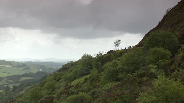 A wide side view of a steep mountainside with a road curving around it midway up, and a large valley with green fields in the background beneath the overcast sky