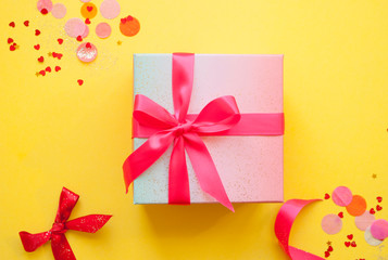 Giftbox tied with pink color ribbon on bright yellow background with sparklets and confetti. Flat lay style.