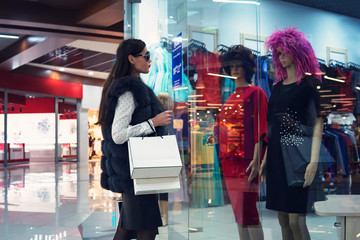 Young woman in shopping centre standing near the shop window and looking at two mannequins in dresses and wigs. Trendy attractive girl is doing shopping in mall. Lady in glasses looking for outfit