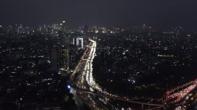 Panoramic night cityscape scene above illuminated downtown city skyscrapers & traffic highways congestion above Tomang, Indonesia. Aerial pan right.