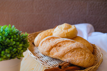 Freshly baked delicious bread in basket on wooden table with white cloth and green plant flowerpot.