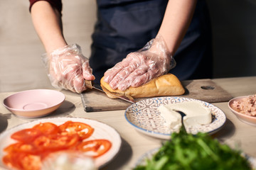 Obraz na płótnie Canvas Focus on hand cutting with knife long bread roll on cutting board for preparing grilled panini sandwich. Ingredients on blurred foreground: tomato slices, tuna, butter, salad on plate at wooden table.
