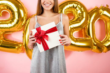 beautiful happy woman holding red gift box in front of 2020 christmas balloons isolated over pink