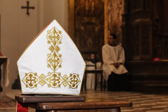 Mitra, copricapo religione Cattolica, appoggiato su un banco durante una cerimonia, chierico sullo sfondo seduto.