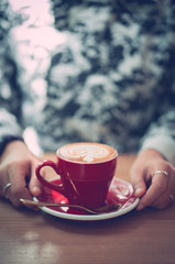 red cup of hot cappuccino coffee with beautiful latte art in blurred woman hands, autumn color tone