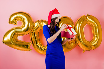 excited beautiful woman wearing santa hat opening gift box in front of new year 2020 balloons isolated over pink