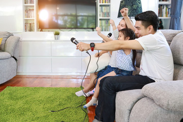 side view happy asian family, father, mother daughter and son singing karaoke on the sofa in the living room with happy smiling face (relaxation and technology concept)