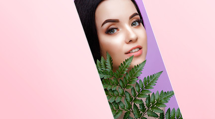 Portrait of young beautiful woman with healthy glow perfect smooth skin holds green tropical leaf. Model with natural nude make up look into the hole of pink paper. Purple background.